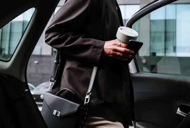 A girl is getting ready to enter the car, wearing a brown coat and a black bag, holding her phone and her cup of coffee